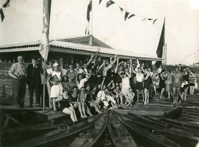 Foto van een groep mensen voor een botenhuis met ervoor houten kano&#039;s
